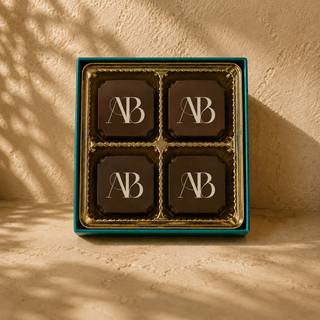 Luxury chocolate gift box featuring four individually wrapped dark chocolates arranged in a gold tray inside a teal box. Each chocolate displays an elegant "AB" monogram in a refined serif style. Warm natural sunlight and soft botanical shadows create a sophisticated, premium presentation against a textured beige background.
