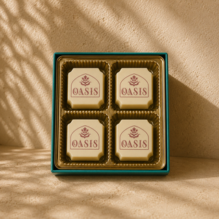 Luxury white chocolate gift box featuring four individually molded chocolates embossed with the Oasis logo, arranged in a gold tray within a rich teal box. Styled in a warm Mediterranean-inspired stucco setting with soft directional sunlight, textured neutral surfaces, and natural shadow play for an elevated editorial product photography look.