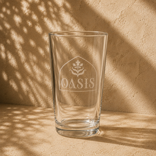 16oz clear glass tumbler featuring an etched Oasis logo with a minimalist botanical emblem, photographed in a warm Mediterranean-inspired stucco setting with soft natural sunlight and textured shadows. Clean modern drinkware design with a refined, premium aesthetic.